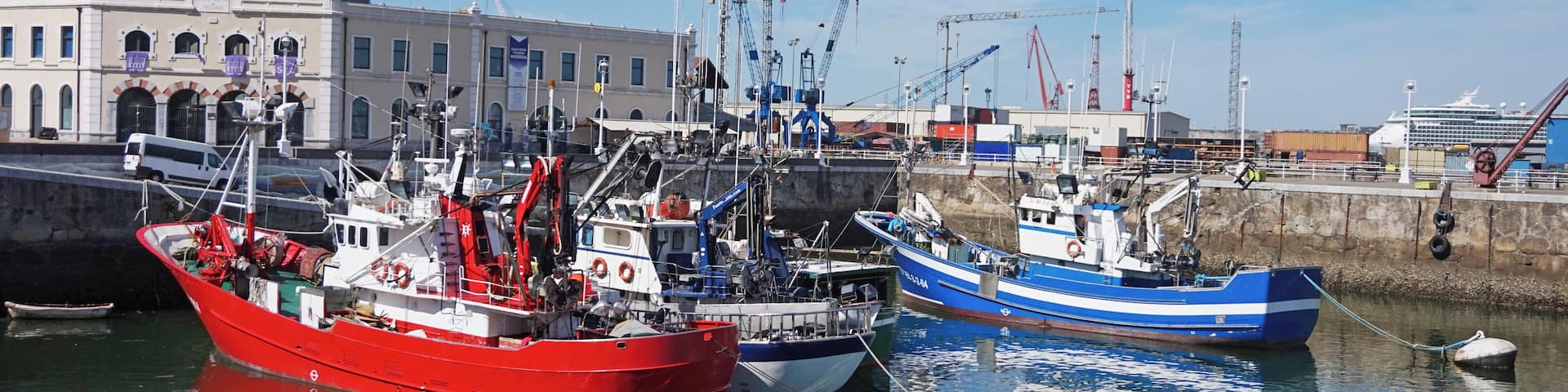 A harbor with several boats, including a red fishing boat and a blue boat, docked near a building with a sign that reads "Marina."
