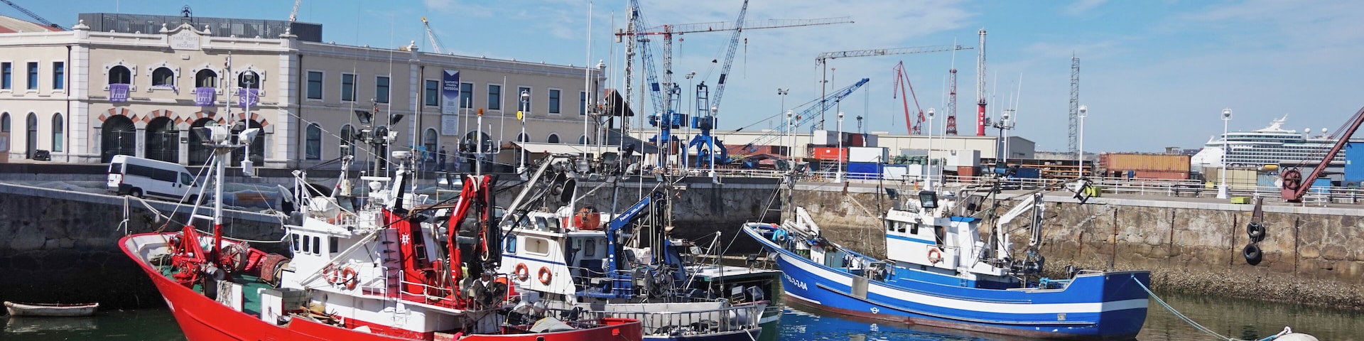 Un puerto con varias embarcaciones, incluyendo un barco pesquero rojo y un bote azul, amarradas cerca de un edificio con un letrero que dice "Marina".