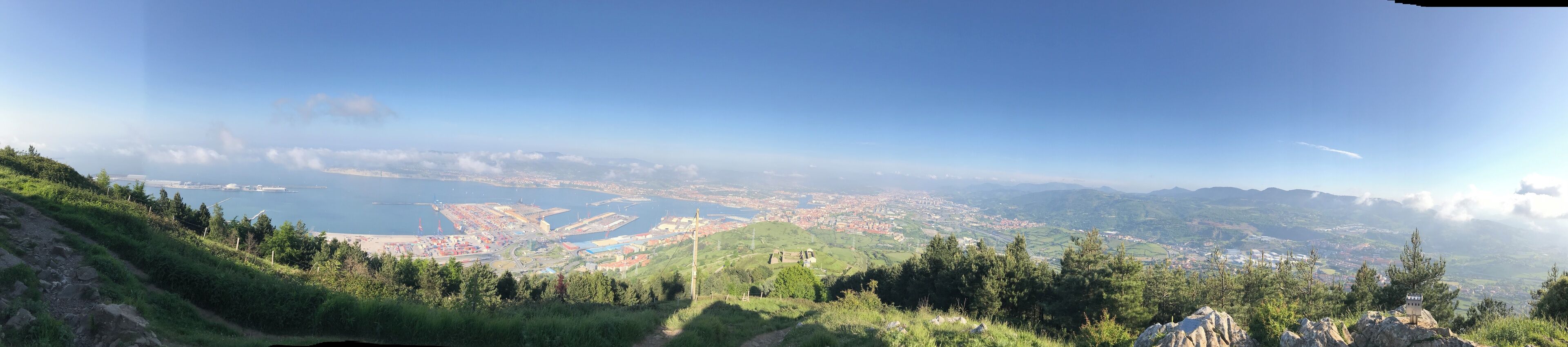 A coastal cityscape with a clear view of the sea, mountains, and a hilly landscape.