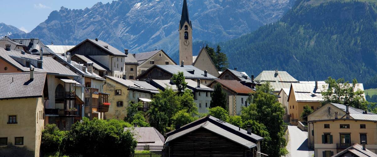 A mountain town with a prominent church tower and snow-capped peaks in the background.