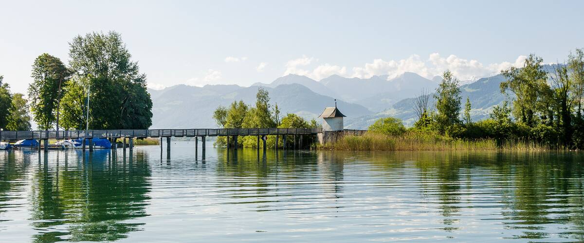 A calm lake with a wooden pier and a small white building, surrounded by lush greenery and mountains in the background.
