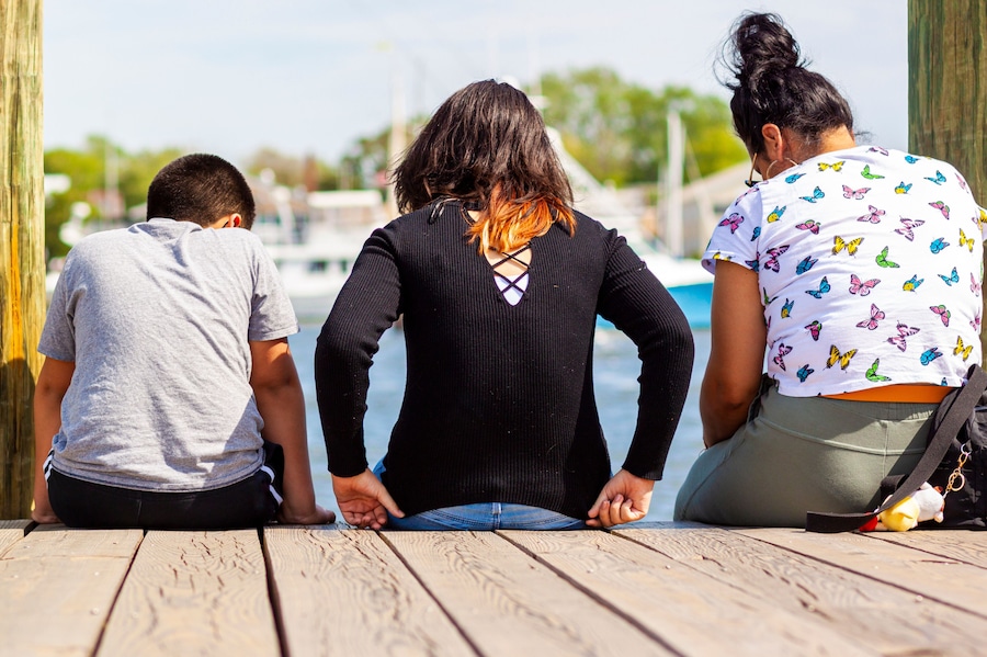 A hispanic family with mother in the middle and her son and daughter on both sides are sitting on the city dock of Annapolis looking at the sea. They are enjoying the beautiful sunny weather.