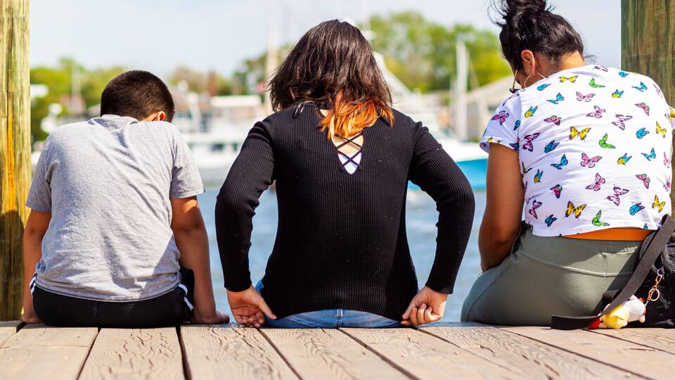 Three people sitting on a wooden dock by a body of water with boats in the background.