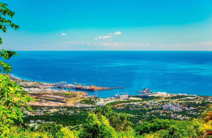 Un paisaje urbano costero con un mar azul cristalino, instalaciones portuarias industriales y abundante vegetación.