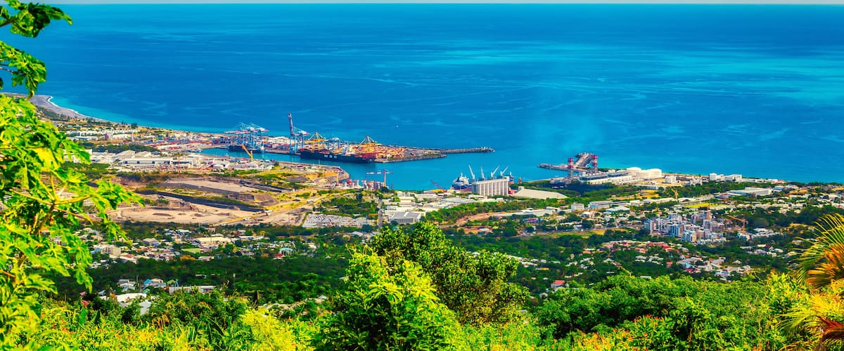 A coastal cityscape with a clear blue sea, industrial port facilities, and lush greenery.