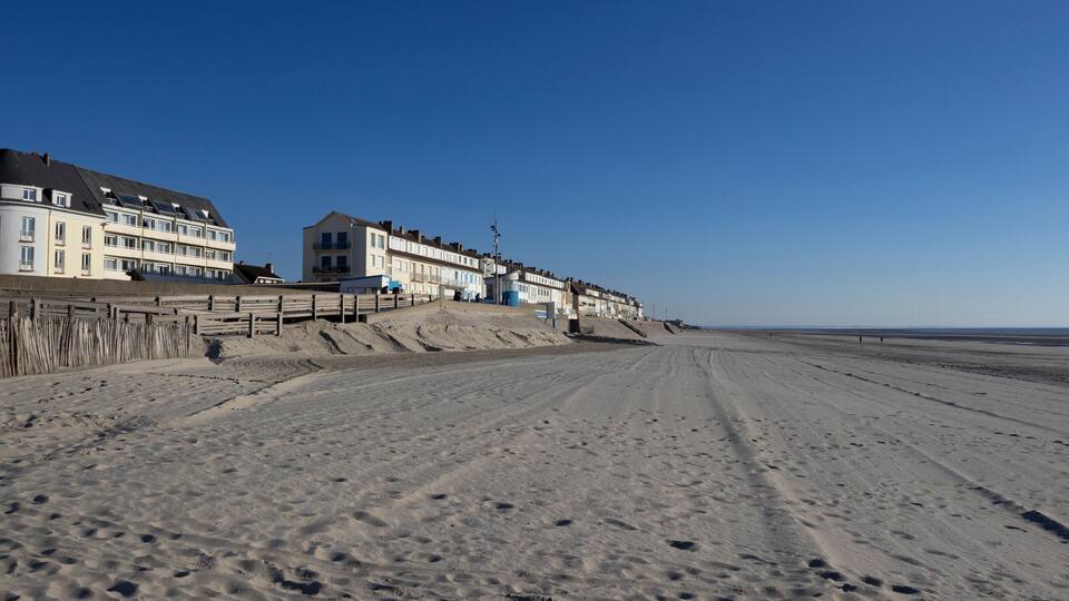 A sandy beach with a wooden boardwalk leading to a row of white buildings with blue doors.