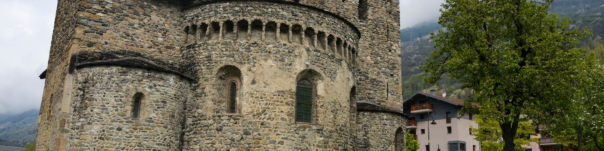 Um edifício histórico de pedra com uma torre, cercado por um muro de pedra e uma área gramada com árvores.