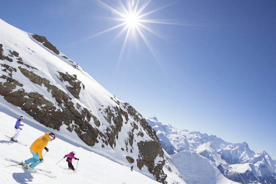 Sciatori su un pendio montuoso innevato, con affioramenti rocciosi e un cielo blu limpido.
