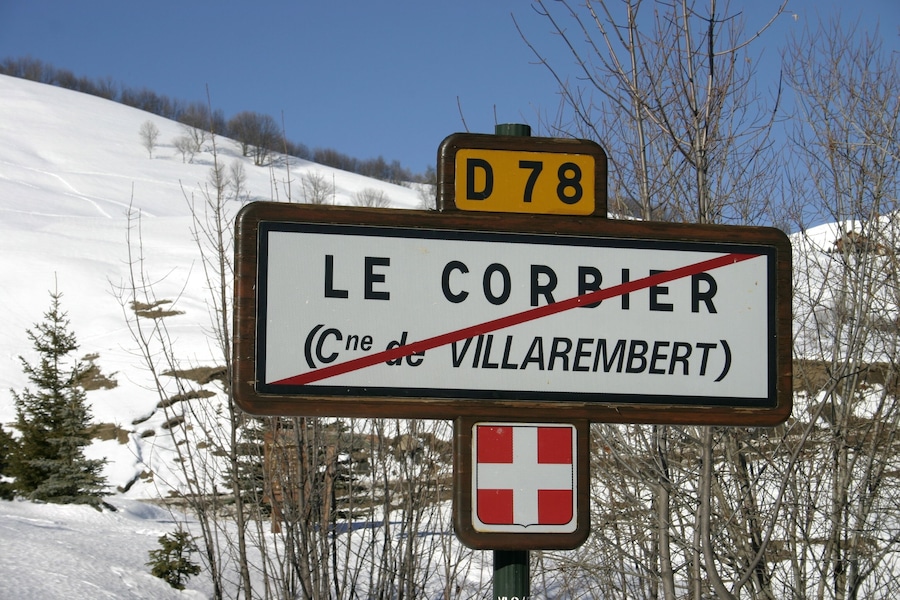 A road sign with 'LE CORBIER' and a red cross symbol, indicating a closed road, with a snow-covered landscape and trees in the background.