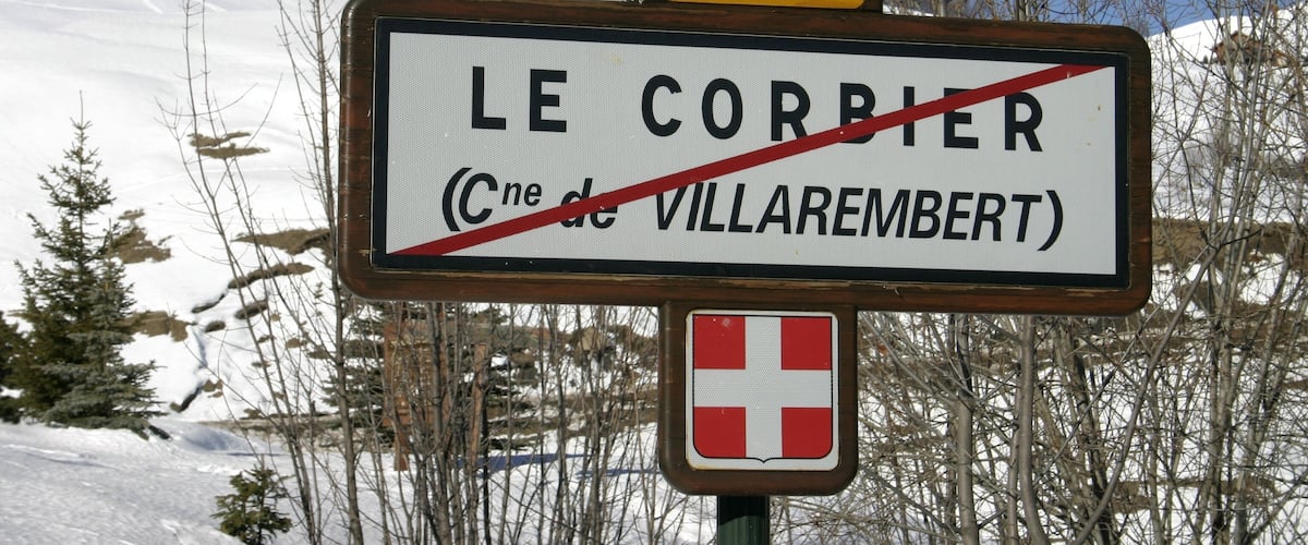 A road sign with 'LE CORBIER' and a red cross symbol, indicating a closed road, with a snow-covered landscape and trees in the background.