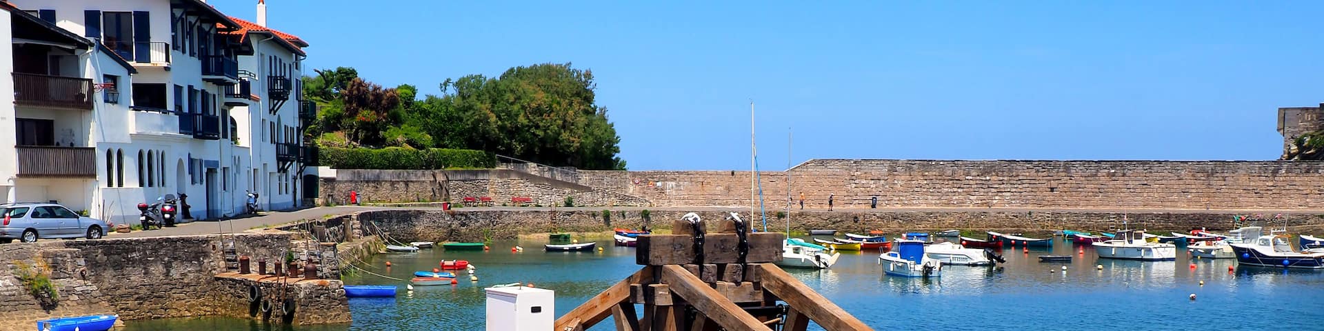 A harbor with several boats docked, a fishing boat with a wooden structure in the foreground, and a building with balconies in the background.