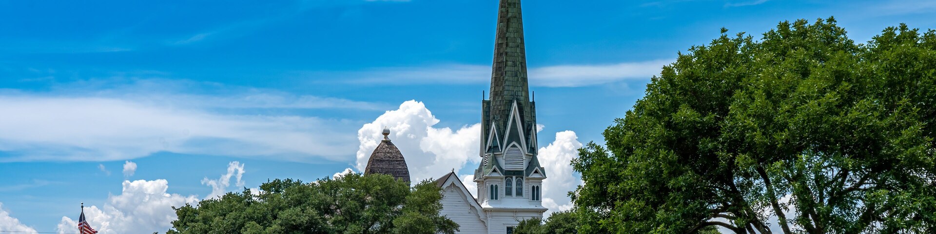 A church with a tall steeple and a cross, surrounded by trees and a clear blue sky.
