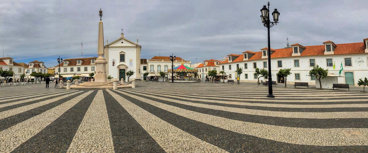 A plaza with a central monument, striped pavement, and surrounding buildings.