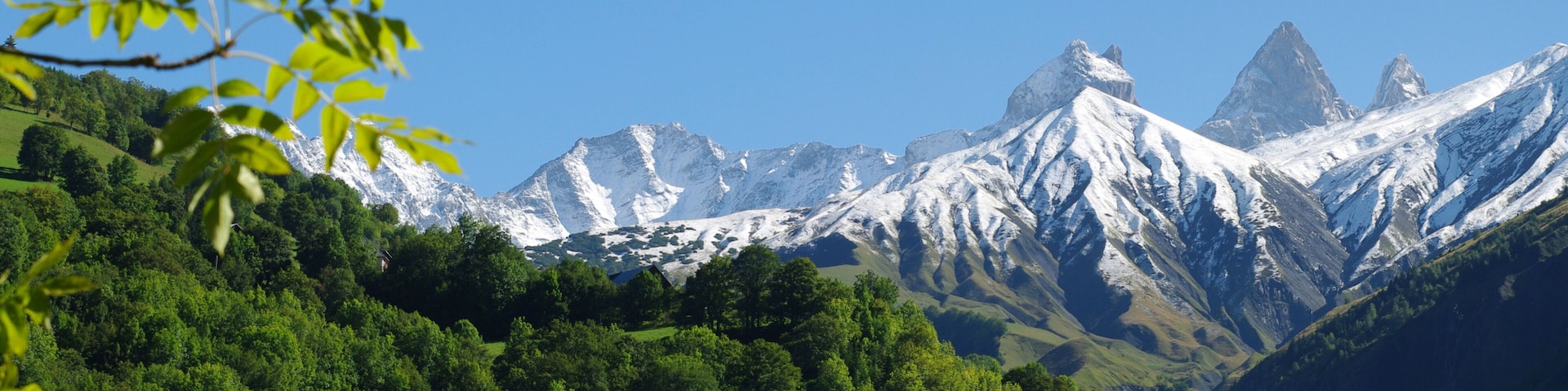 Paisagem montanhosa com picos nevados, floresta verde densa e céu azul límpido.
