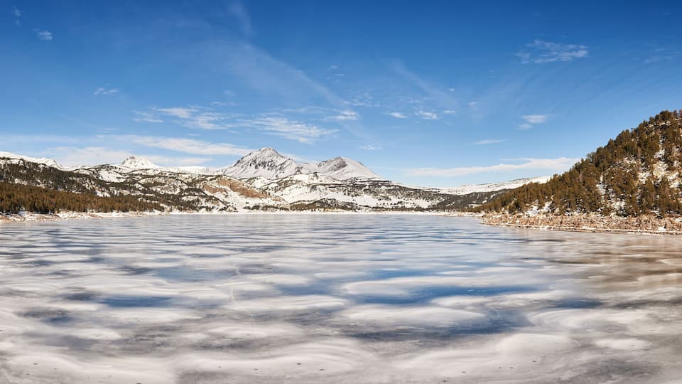 A frozen lake surrounded by snow-covered mountains and dense forests.