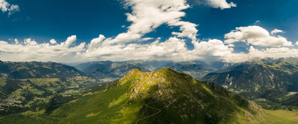 A mountain range with a prominent peak, lush green valleys, and a clear blue sky with scattered clouds.