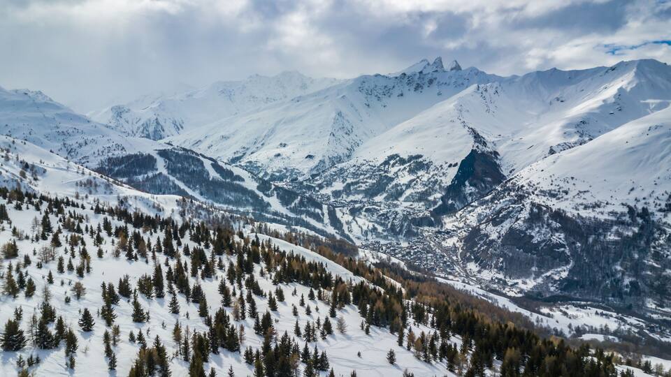 A snow-covered mountain range with a valley and scattered trees.