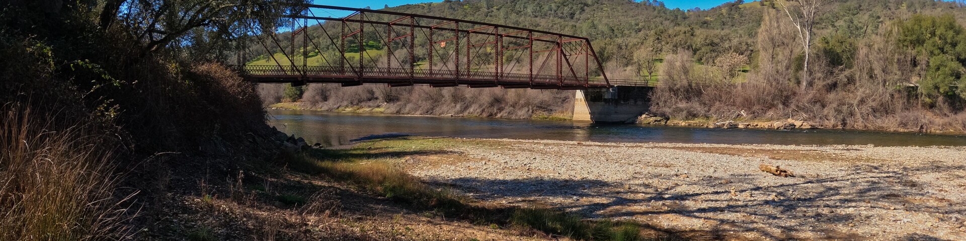 Un puente de metal cruza un río, rodeado de árboles y colinas.