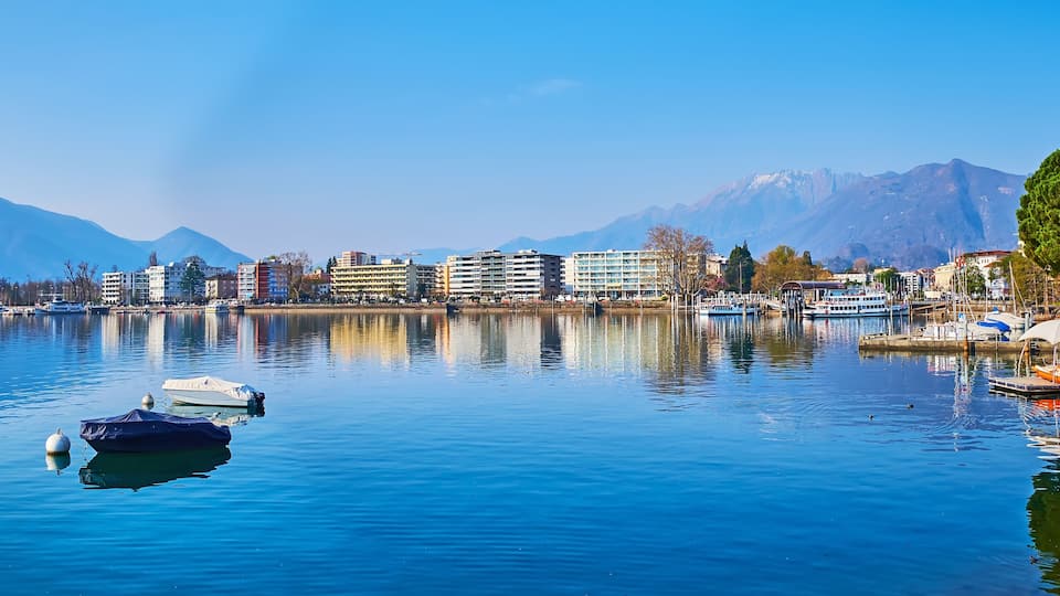 A calm lake with boats, buildings, and mountains in the background.
