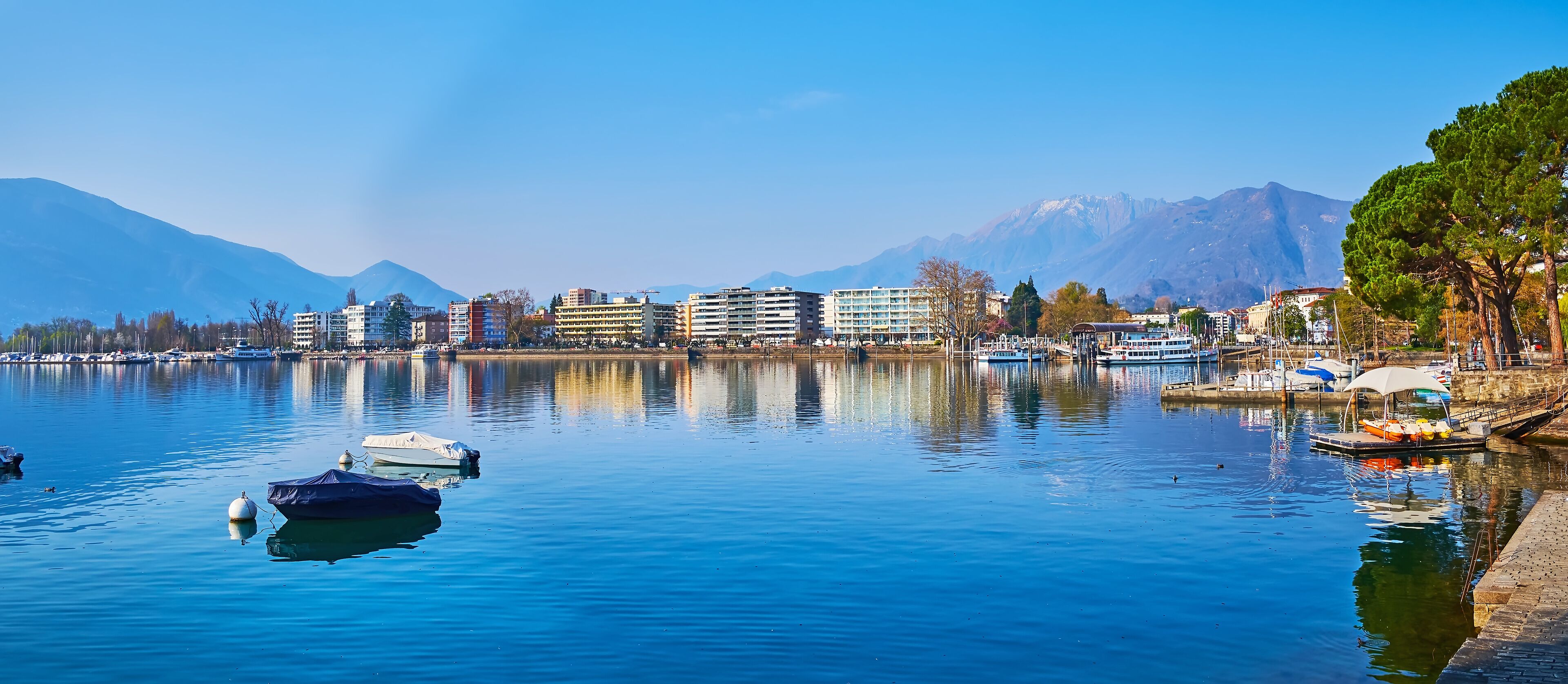 A calm lake with boats, buildings, and mountains in the background.