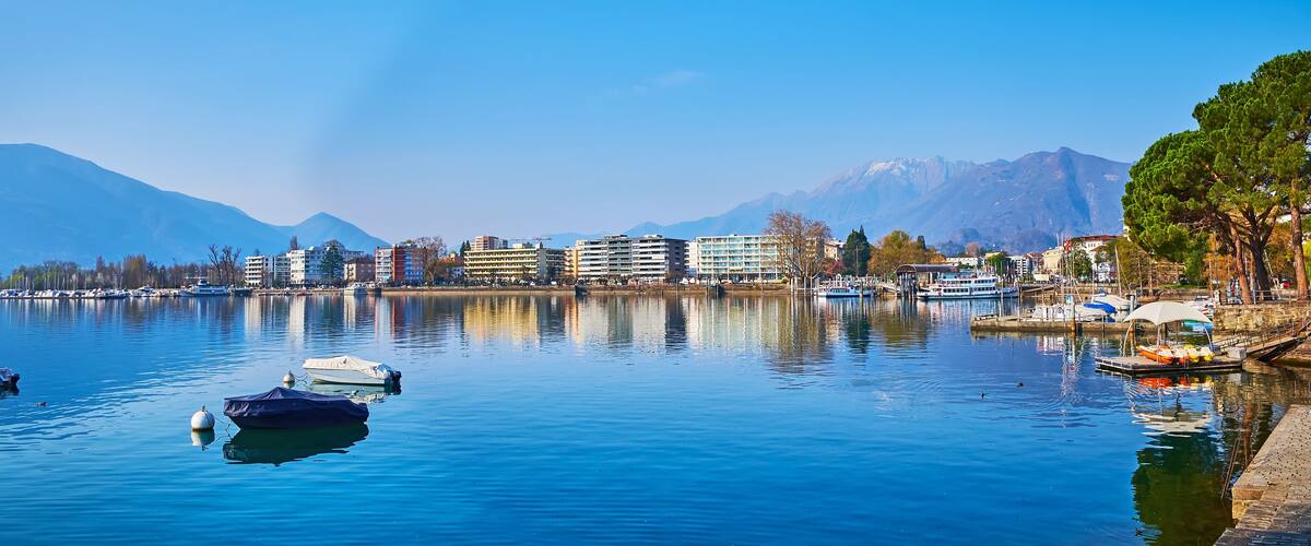 A calm lake with boats, buildings, and mountains in the background.