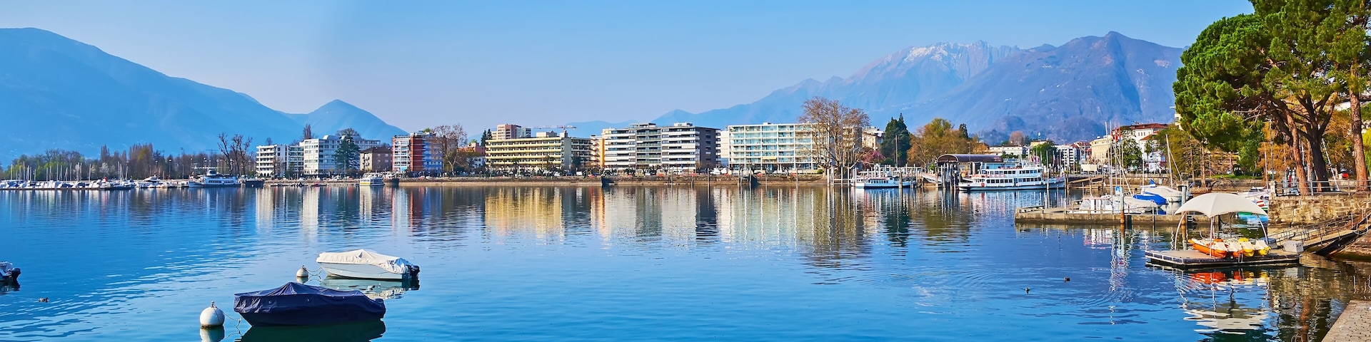 A calm lake with boats, buildings, and mountains in the background.