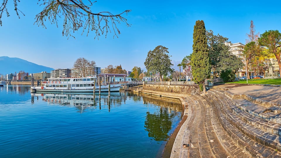 A calm lake with boats, a stone embankment, and buildings in the background.