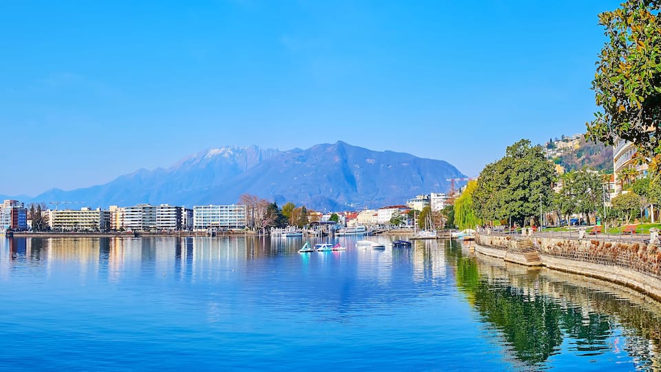A calm lake with boats, a cityscape, and mountains in the background.