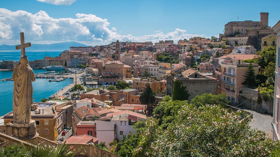 A coastal town with historic buildings, a prominent statue, and a castle on a hill.