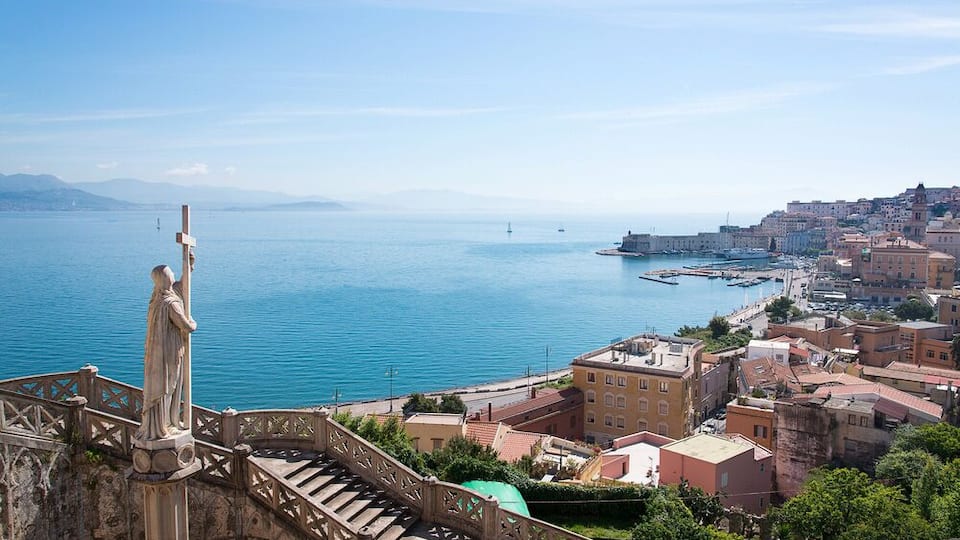 A coastal city with a prominent statue, a staircase, and a harbor with boats.