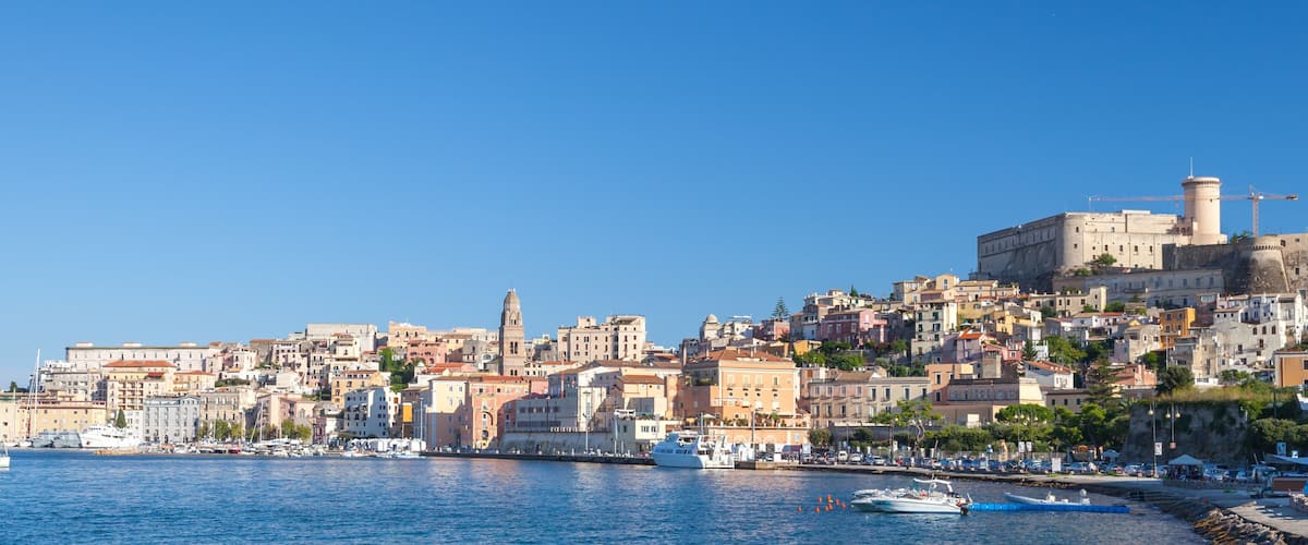 A coastal cityscape with historic buildings, a prominent tower, and boats in the harbor.