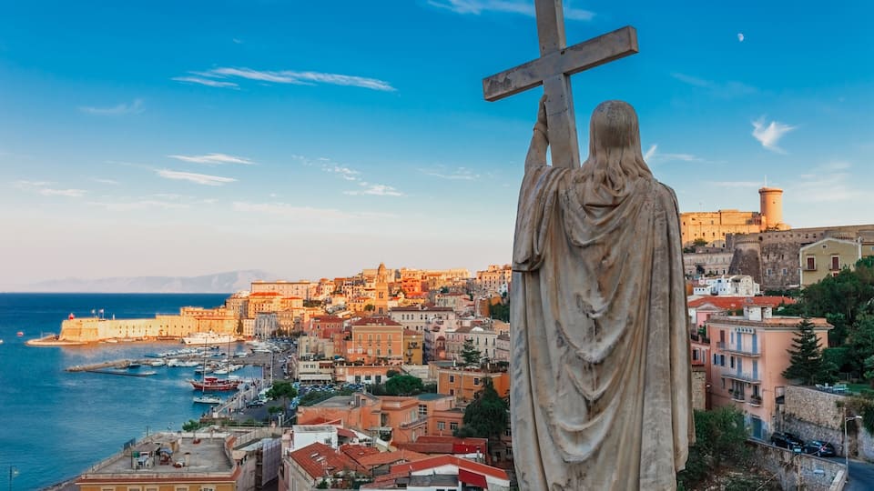 A statue with a cross, overlooking a coastal town with historic buildings and a harbor.