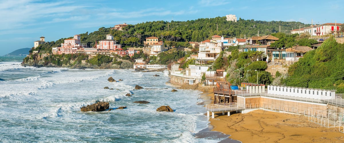 A coastal town with buildings on a hillside, waves crashing onto the shore, and a sandy beach.