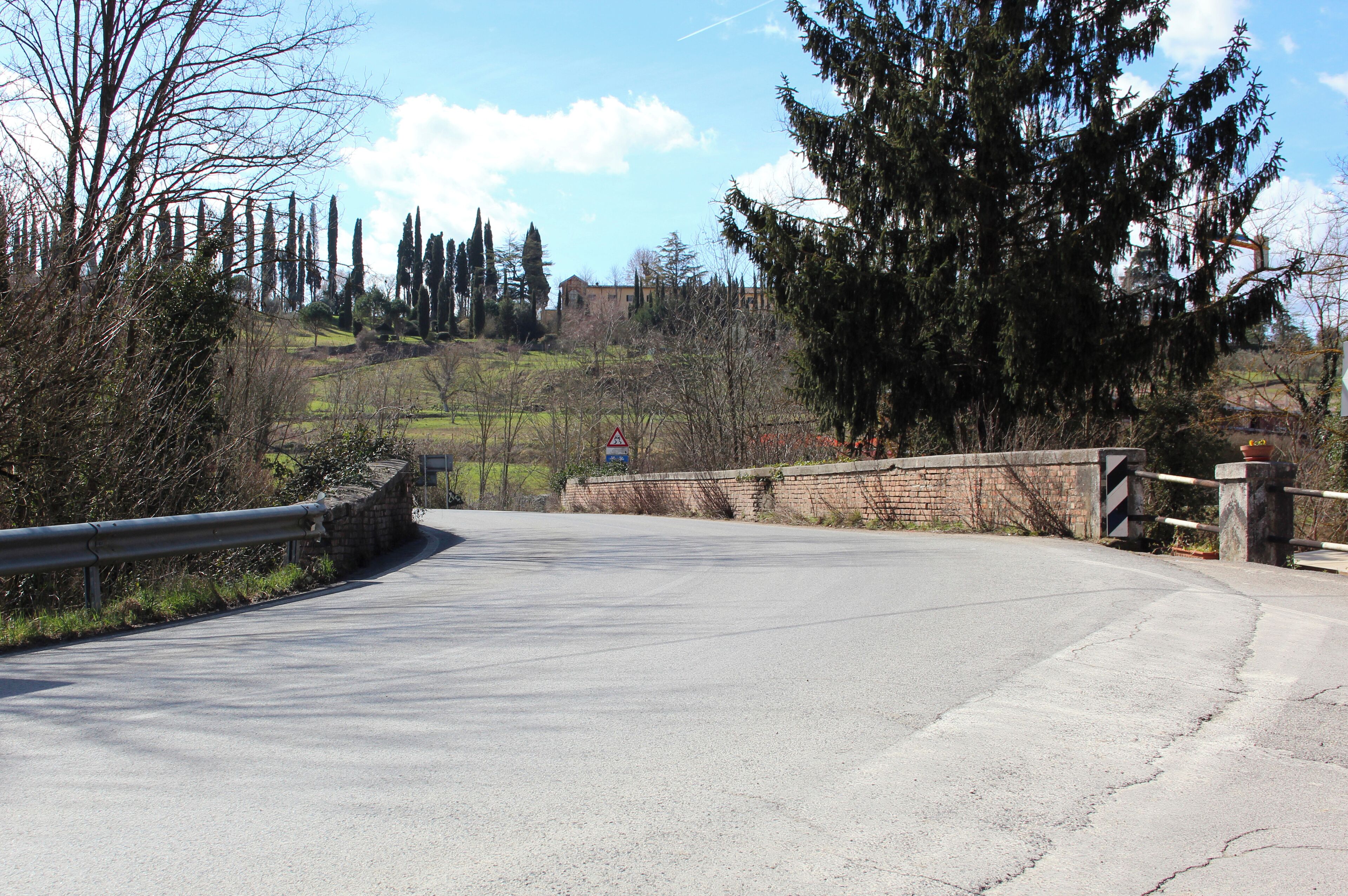 A winding road with a stone bridge, surrounded by trees and a distant building.
