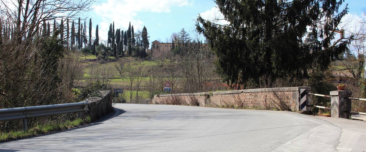 A winding road with a stone bridge, surrounded by trees and a distant building.