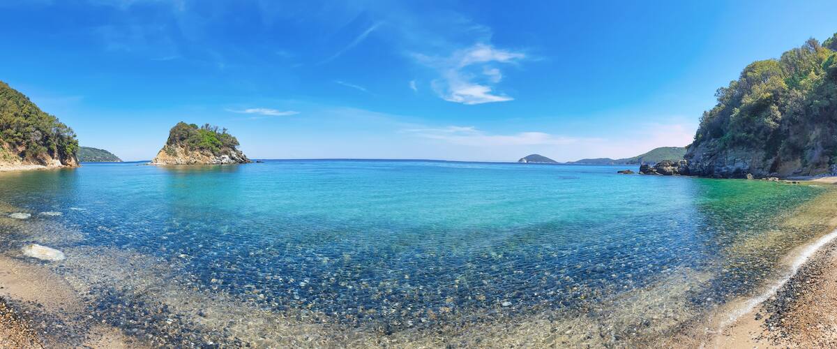 A clear coastal landscape with a pebble beach, calm turquoise waters, and green hills.