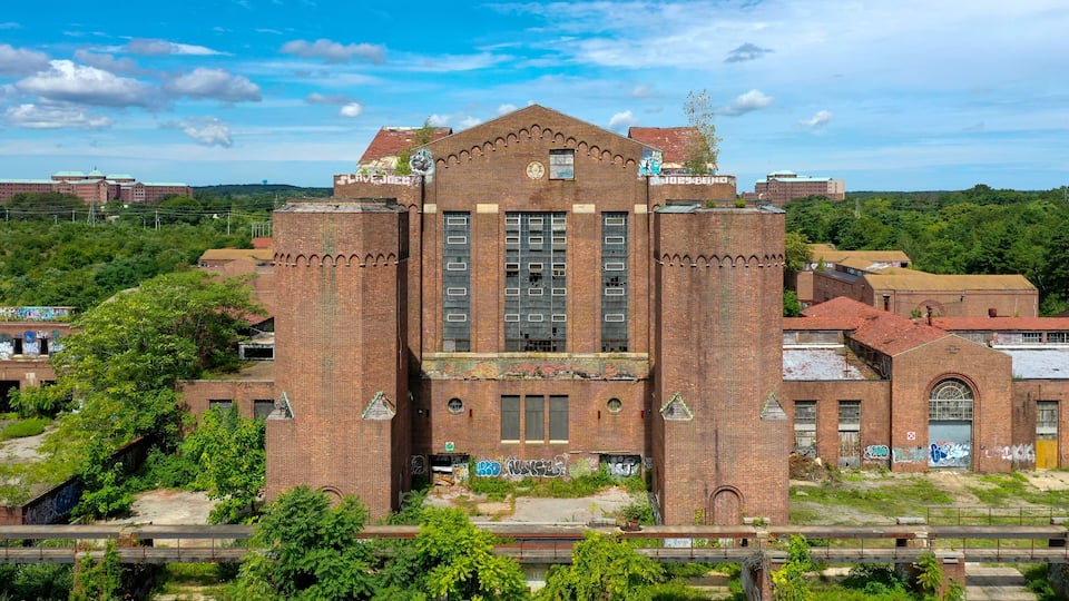 A large, abandoned brick building with graffiti, surrounded by greenery and a railway track.