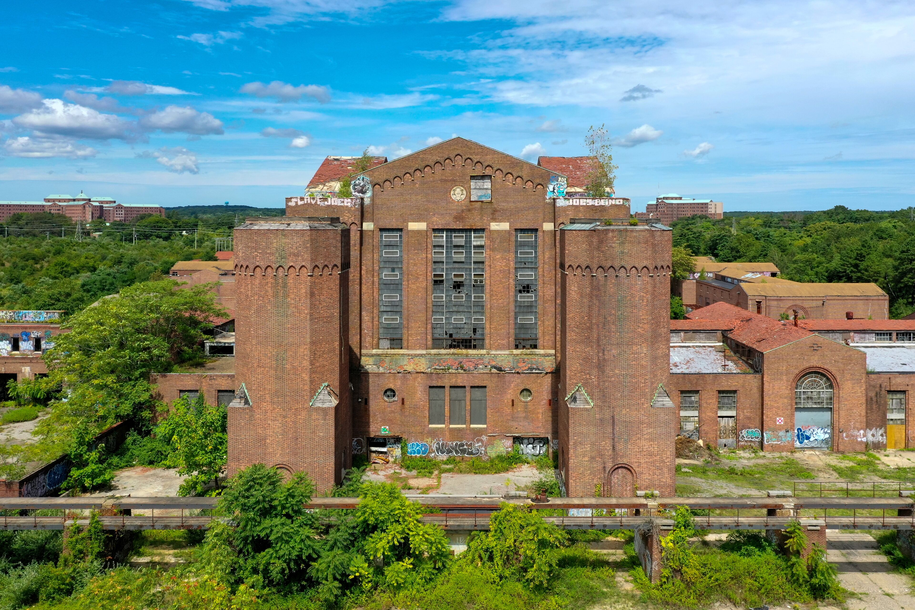 A large, abandoned brick building with graffiti, surrounded by greenery and a railway track.