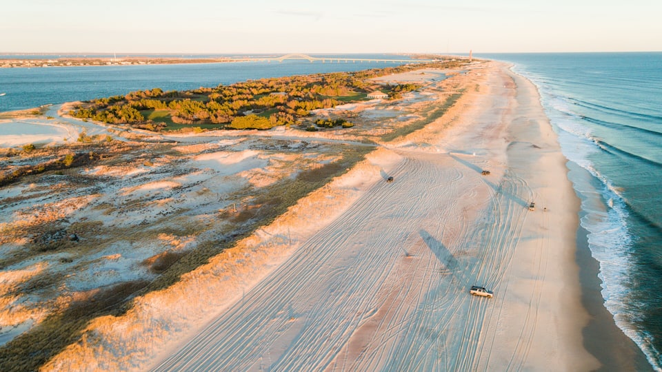 A coastal landscape with a sandy beach, a dune system, and a bridge in the distance.