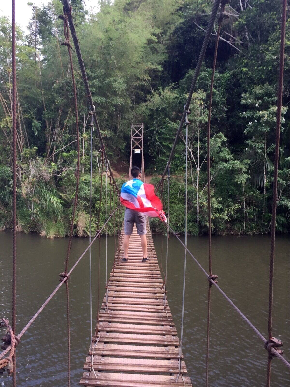 A person crossing a wooden suspension bridge over a river, surrounded by dense greenery.