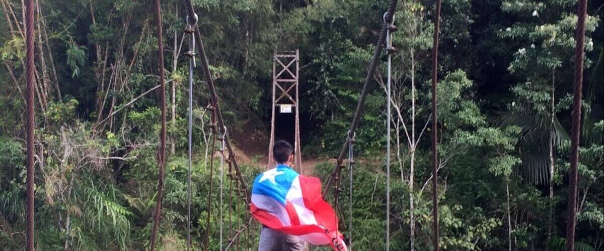 A person crossing a wooden suspension bridge over a river, surrounded by dense greenery.