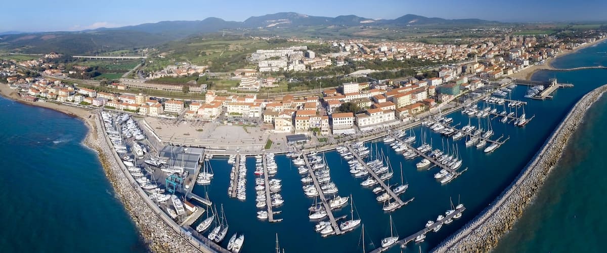 An aerial view of a coastal town with a marina, buildings, and surrounding hills.