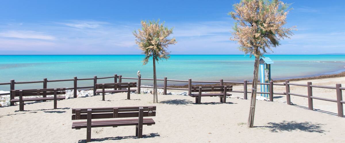 A sandy beach with wooden benches and a wooden fence, overlooking a clear blue sea.