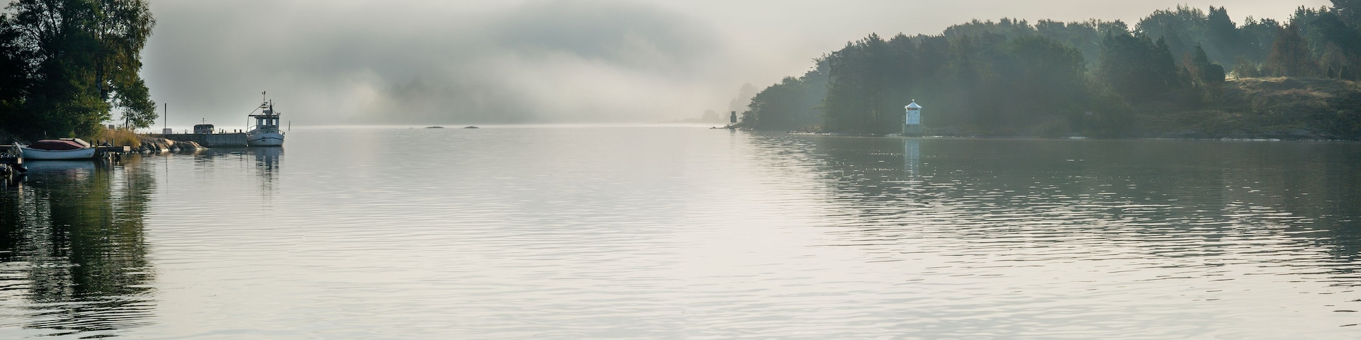 Un lago tranquilo con un bote amarrado a la izquierda, un ave volando en la distancia y un faro a la derecha.