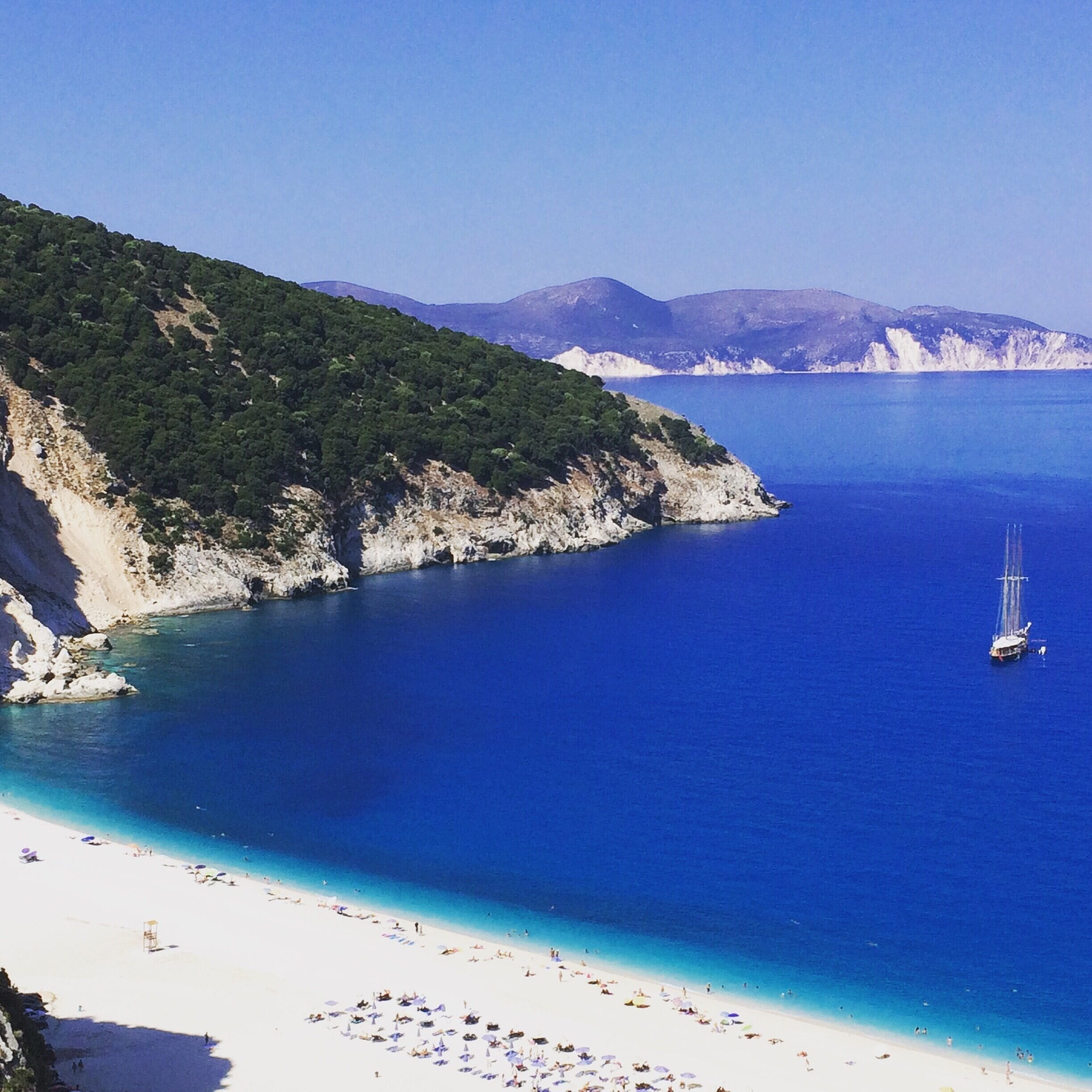 A coastal landscape with a clear blue sea, a sandy beach, and a sailboat.
