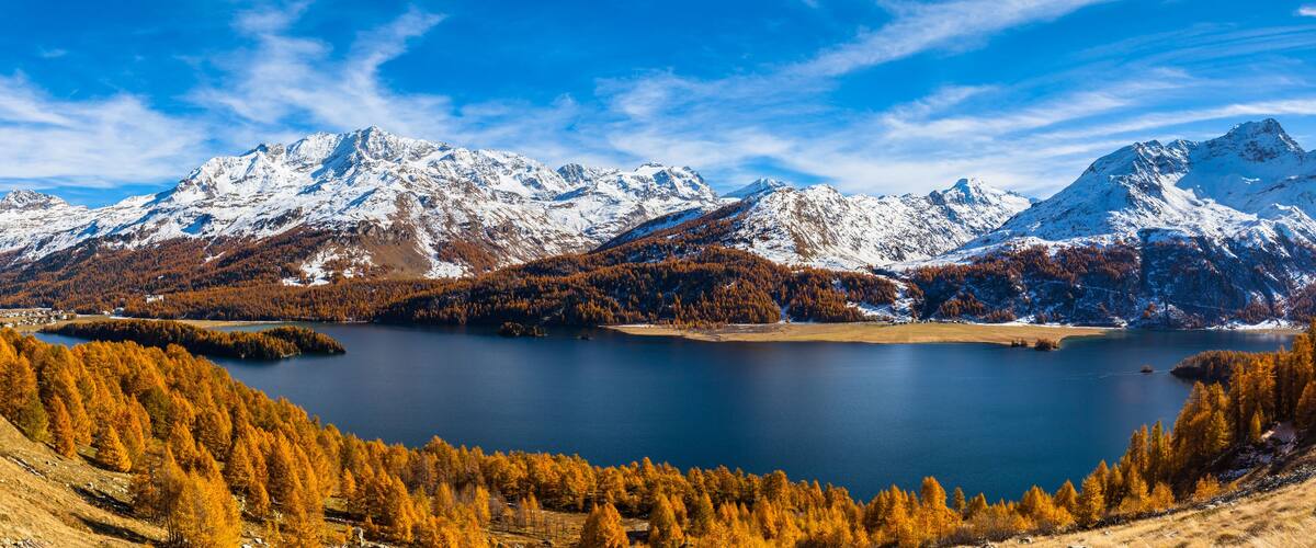 A mountainous landscape with a lake surrounded by autumn-colored trees and snow-capped peaks.