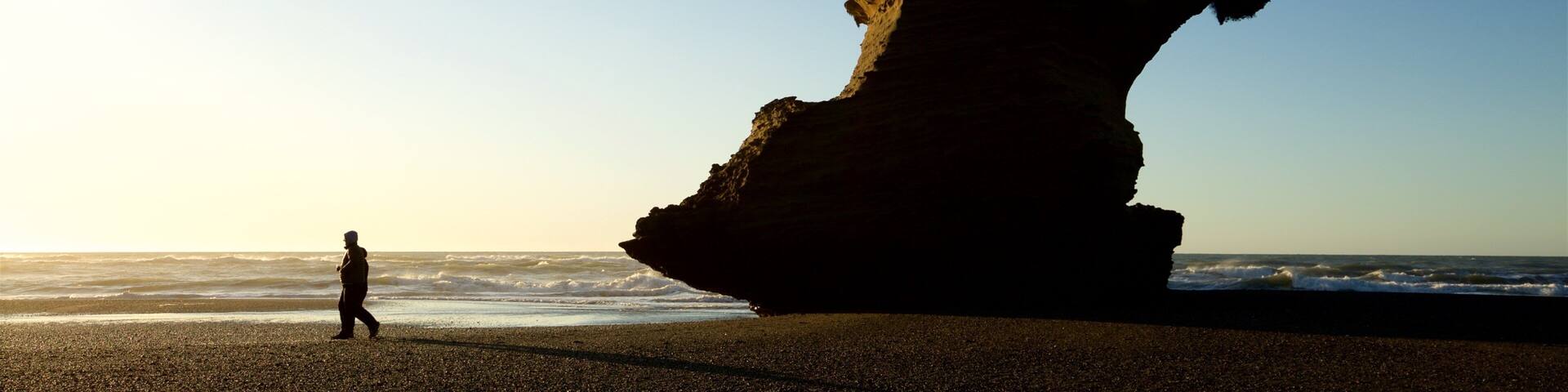 A person walking on a beach with a large rock formation in the background.