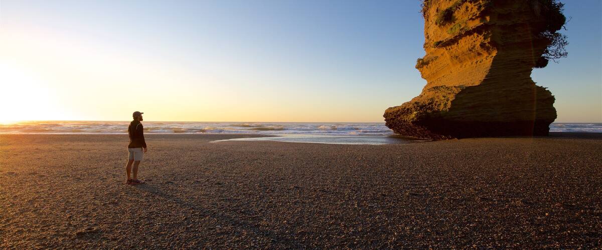 A person standing on a pebbled beach with a large rock formation in the background.
