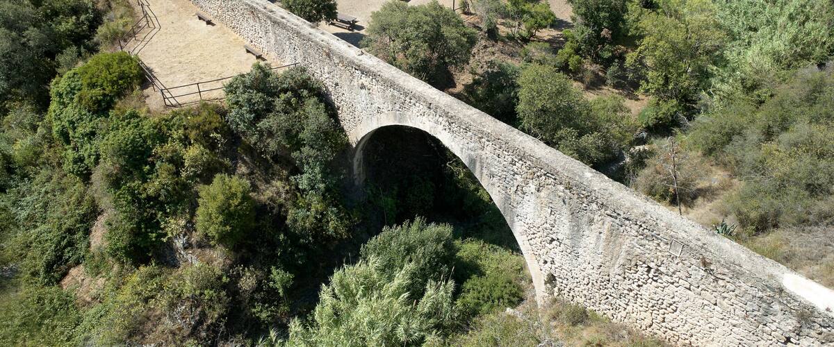 A stone bridge over a dry riverbed, surrounded by dense greenery and trees.