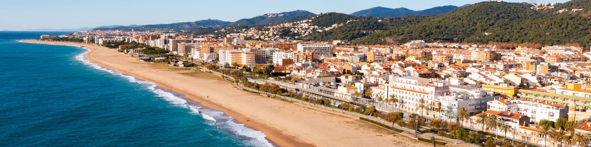 A coastal city with a long sandy beach, dense urban area, and green hills in the background.